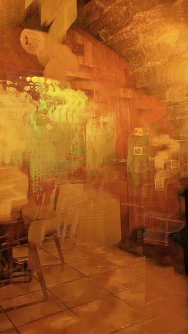 A double exposure or motion-blurred photograph of an indoor space, possibly a cafe or restaurant, rendered in warm orange, yellow, and green hues.