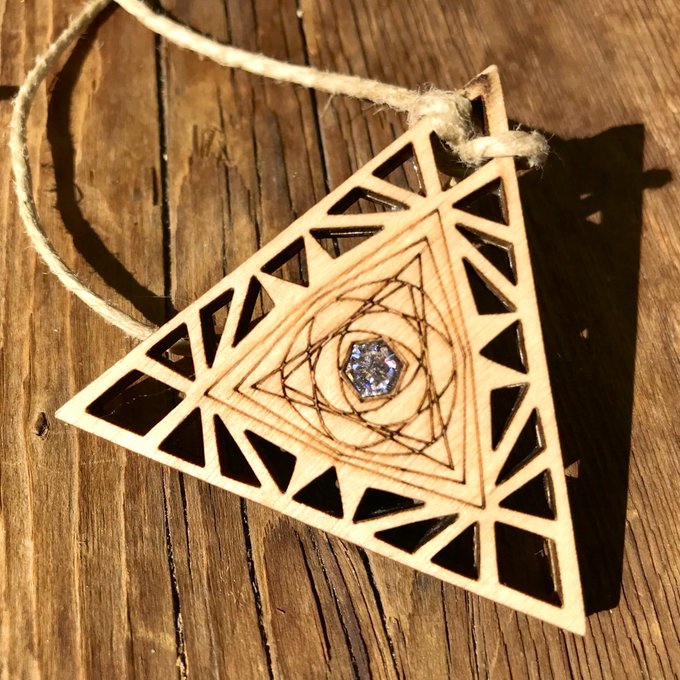 A triangular wooden pendant with geometric cutouts and a small blue gemstone hangs from twine on a weathered wooden surface.