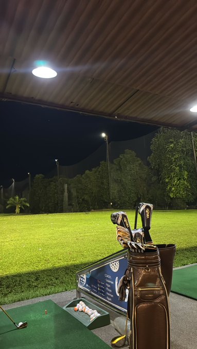 A nighttime view of a covered golf driving range with golf clubs in a bag in the foreground and illuminated balls lined up on the mat.