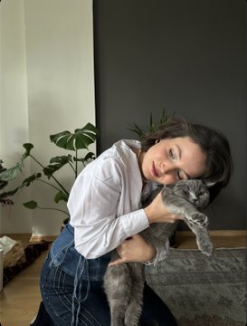 A woman in a white shirt and jeans hugs a gray cat in a cozy indoor setting with green plants in the background.