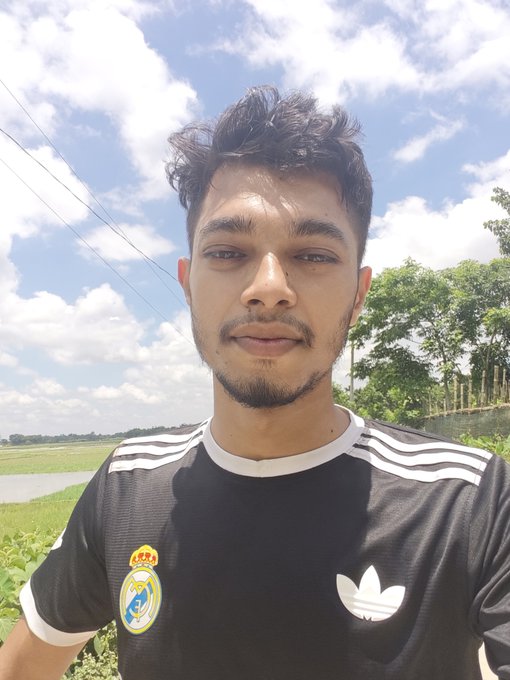 A young man with a goatee wearing a black Adidas t-shirt takes an outdoor selfie with a blue sky and trees in the background.