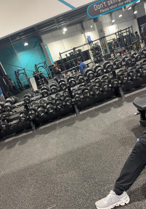 A gym with a blue wall and white ceiling features black dumbbells on racks, with two individuals present.