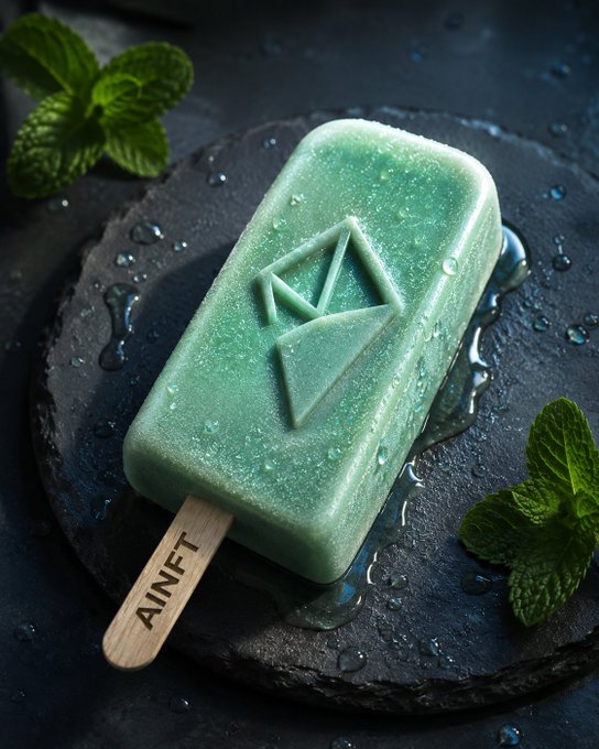 A green popsicle with a geometric logo embossed on it sits on a slate plate garnished with fresh mint leaves, its wooden stick stamped with the text "AINFT," visually representing the brand behind the AINFT Nova platform mentioned in the post.
