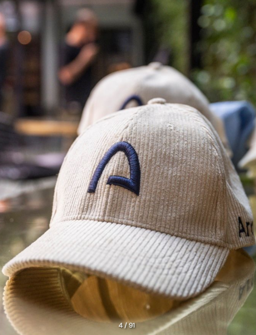 A close-up photo of a beige corduroy baseball cap with a dark embroidered logo, resting on a reflective surface with a blurred background.