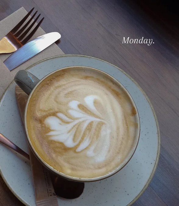 The image features a cup of coffee with white foam on top, placed on a plate next to a fork and knife. The text "MONDAY" can be seen at the bottom right corner of the photo. The background appears to be a wooden table or countertop, which could potentially be part of a restaurant setting.