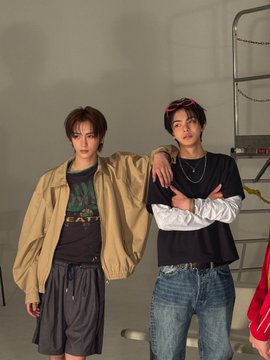 The image features two young men standing next to each other, both wearing jeans and jackets. One of them has a black shirt with white lettering, while the other wears a tan jacket over his blue jeans. They are positioned against a gray wall, which provides a neutral backdrop for their attire. The post they appear in is titled "#ALPHADRIVEONE #ALD1 #알파드라이브원 같은 경우", and it seems to be a social media platform where people share their outfits or fashion-related content.
