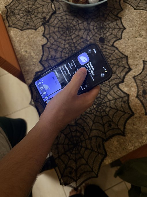 A hand holds a smartphone displaying the Phantom crypto wallet interface over a Halloween-themed spider web placemat, as the user prepares to onboard their little brother into the world of cryptocurrency.