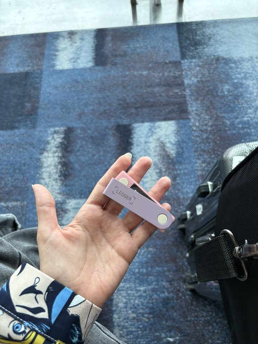 A hand holds a purple Ledger crypto hardware wallet, with the "LEDGER" logo visible, set against a blue checkered carpet near airport luggage. The image humorously illustrates the social media caption about being stopped at the Hong Kong airport by a curious customs officer who mistook the crypto wallet for just a "cute USB," while the traveler nervously held their life savings.