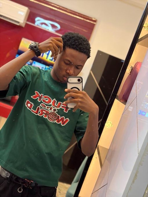 A young man taking a mirror selfie while wearing a green t-shirt that reads 'FUCK THE WORLD' in red text.