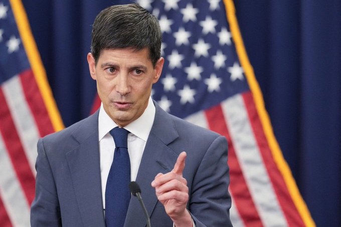 A man in a dark suit speaking at a podium with American flags in the background.