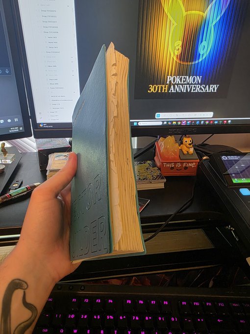 A hand holds a light blue book with worn, yellowed page edges embossed with 'HOLOFOLDER' (likely 'FOLKOLDER') in front of a gaming setup featuring dual monitors, with a 'Pokémon 30th Anniversary' display visible in the background.