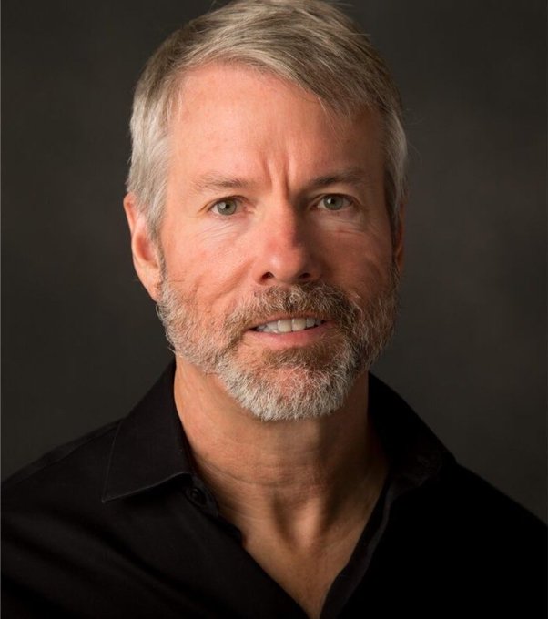 A professional studio portrait of an older man with grey hair and a short beard, wearing a black button-down shirt against a dark grey background.
