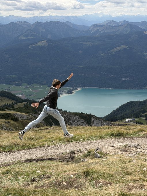A man leaps joyfully across a grassy mountain ridge with a turquoise lake and alpine peaks visible in the valley below.