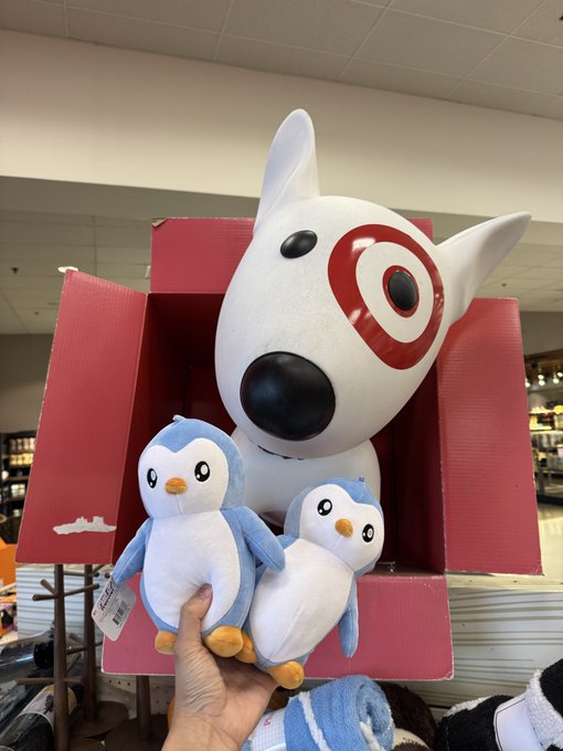 A hand holds up two small blue and white penguin plushies in front of a much larger white dog/bull terrier plush with a red target logo, displayed in a large red box inside a Target store. The image showcases the "Pudgy Penguins" plush toys being held next to what appears to be Bullseye, Target's iconic mascot dog, highlighting the collaboration's location.