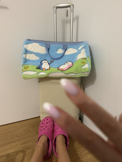 A fluffy travel bag featuring cartoon penguins rests on top of a beige rolling suitcase, with someone wearing pink Crocs and a peace sign gesture visible in the foreground. The cute penguin-themed luggage perfectly complements the "Pengu morning & gm ✈︎" travel-ready vibe.
