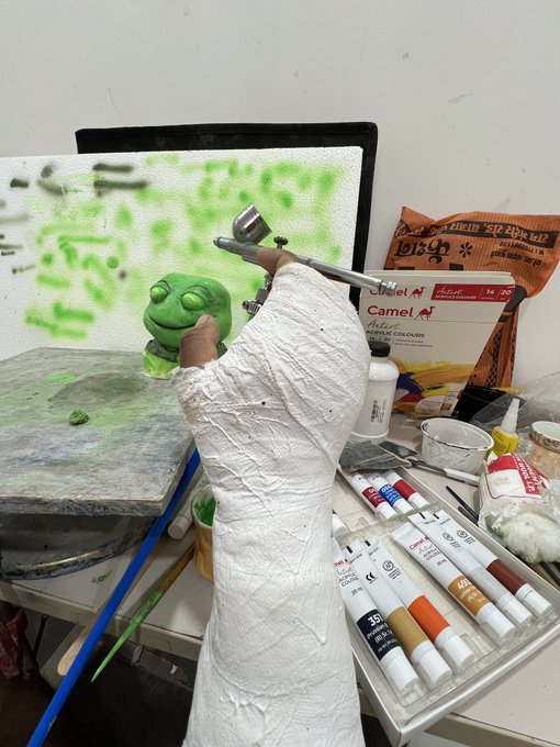 A sculpted white hand holds a green frog figure in an artist's workspace filled with Camel acrylic colors and paint tubes, capturing the creative process behind the "$TOAD" piece. The in-progress sculpture stands ready for painting alongside various art supplies.