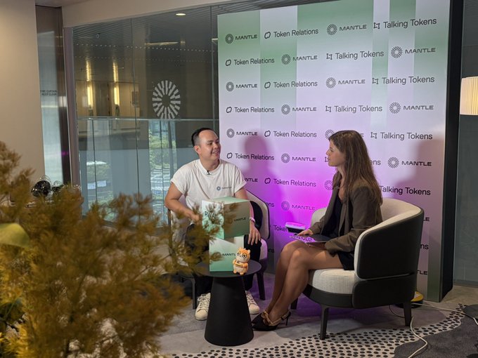 Two people sit in chairs during an interview setup, with a backdrop displaying 'MANTLE', 'Token Relations', and 'Talking Tokens' logos.