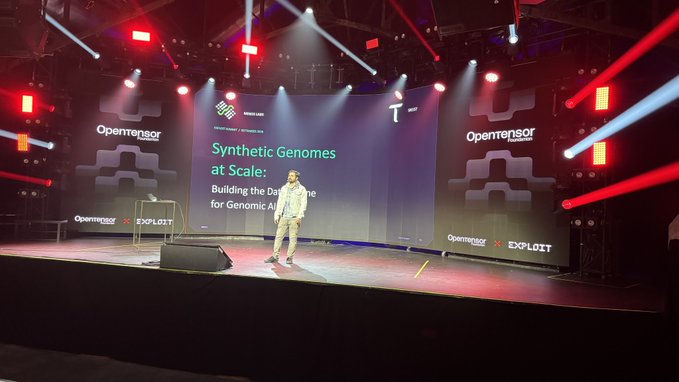A presenter stands on a stage with red and white lighting. A large screen displays 'Synthetic Genomes at Scale: Building the Da... for Genomic AI', with 'Opensor' and 'EXPLOIT' logos visible on the stage backdrop.