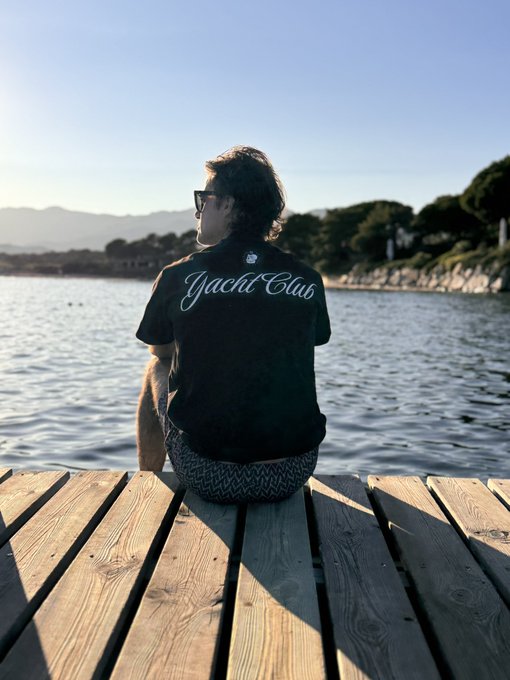 A person wearing sunglasses and a black 'Yacht Club' t-shirt sits on a wooden dock overlooking water and mountains at sunset.