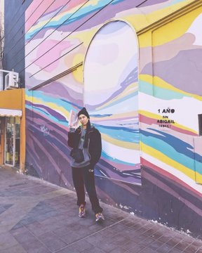 A person in a black hoodie, beanie, and sunglasses standing in front of a colorful striped mural on a city street.