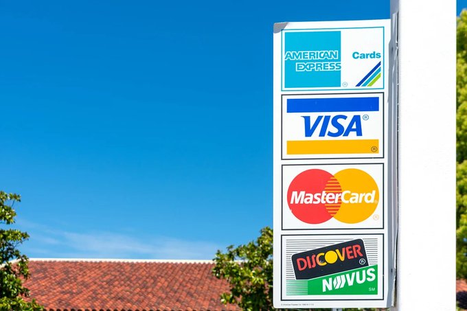 A roadside sign displaying logos for American Express, Visa, MasterCard, and Discover/Novus credit cards against a blue sky.