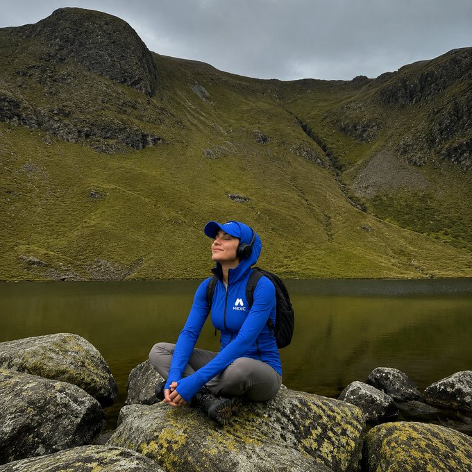 A person wearing a blue MEXC branded jacket and cap sits on rocks by a lake, enjoying the mountain scenery. This image visually represents the "touch grass" and "a little bit of both" options from the accompanying post.
