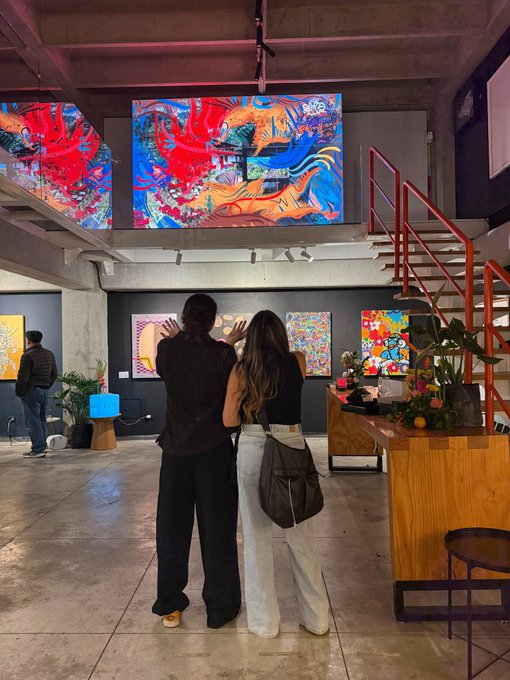 Two people viewed from behind looking up at a large, colorful digital art display of a dragon in a modern art gallery with concrete walls and an orange staircase.