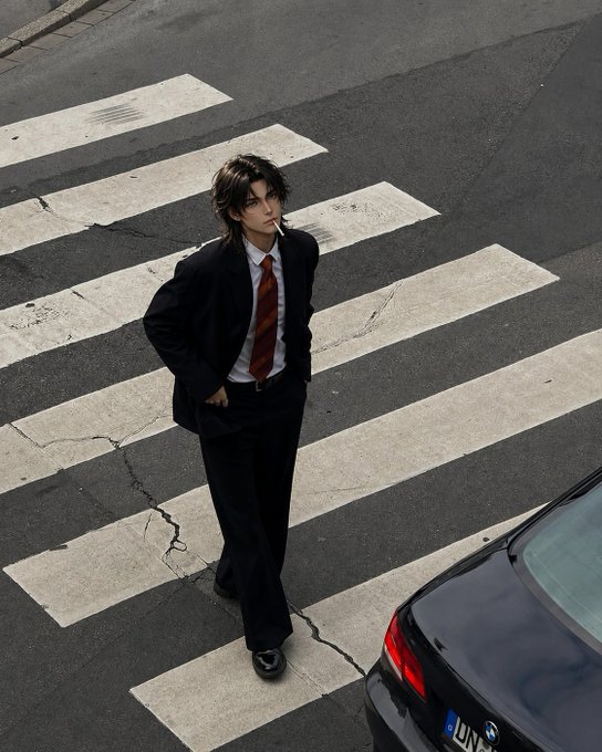 A young man with messy dark hair, wearing a dark suit and striped tie, stands smoking on a crosswalk with a cigarette in his mouth, with a black BMW partially visible on the right.
