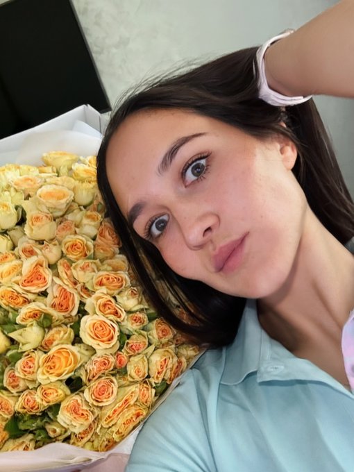 A selfie of a woman with long dark hair posing next to a large bouquet of peach and cream-colored roses.