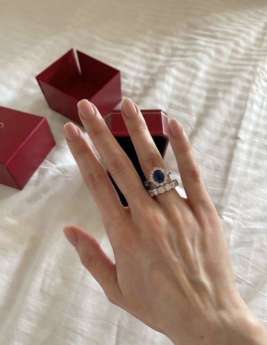 A close-up of a hand displaying a blue sapphire and diamond ring, with two red jewelry boxes resting on a white fabric background.
