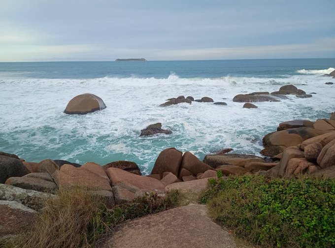 A scenic coastal view shows waves crashing against large brown boulders under a cloudy sky, with a small island visible on the horizon.