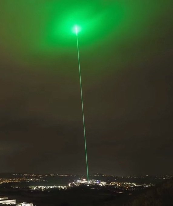 A dramatic nighttime cityscape illuminated by a powerful green laser beam shooting straight into the sky, evoking the hype of a "moonshot" announcement. The intense, singular beam symbolizes the bold promise of a memecoin "dropping" imminently, reinforcing the post's urgent call not to miss the launch.