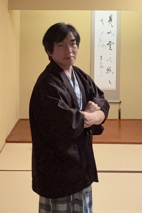 A man wearing glasses and a traditional Japanese black kimono over a patterned yukata, standing with arms crossed in a tatami room with a hanging scroll calligraphy artwork behind him.