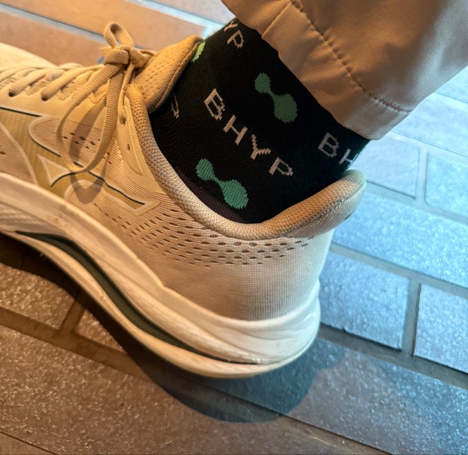 A close-up of a person's foot wearing a white Mizuno running shoe and black socks with green dumbbell patterns and the letters 'BHYP'.