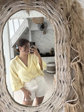 A woman in a yellow blouse and white shorts takes a mirror selfie in a rustic woven-framed oval mirror. The fresh, light-colored outfit complements the uplifting "new week, new opportunities" sentiment, reflecting a bright and confident start to the week.