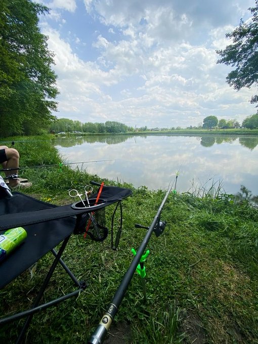 The image shows a person sitting in a chair by a lake, enjoying their time fishing. The scene includes a chair with a backpack next to them, as well as various fishing equipment such as a rod, line, and tackle box nearby. There is also a bottle of soda placed on the ground near the chair. The overall atmosphere suggests relaxation and leisurely outdoor activities like fishing by the lake.