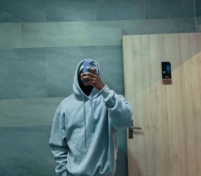 The image features a man wearing a blue hoodie taking a selfie with his cell phone held up to his face against a tiled wall backdrop. The photo captures him from behind, giving an interesting perspective of himself as he poses for the picture.