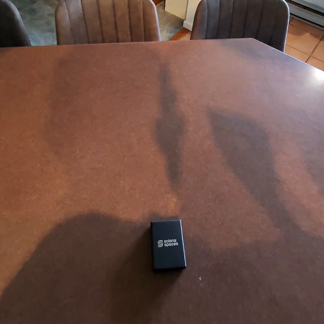 The image features a brown wooden table with a small black box placed on top of it. The text in the image reads "Solana heritage pendant just touched down! such a sick piece from @solarpaces if you don' t already own solana merch, go to their site right now." This suggests that someone has recently purchased or received this item and is sharing information about it with others on social media.
