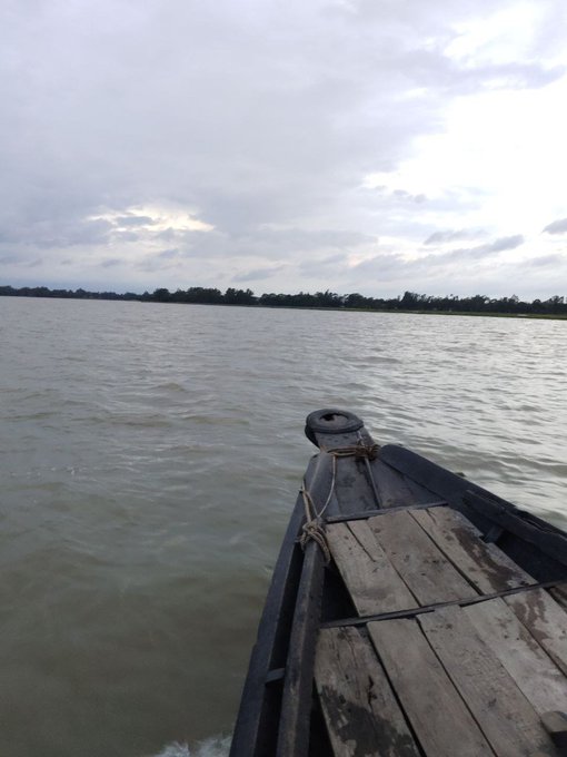 A view from a wooden boat on a body of water under a cloudy sky, with a distant shoreline visible ahead.