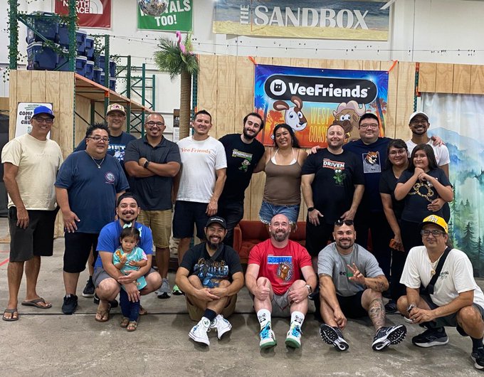 A group photo of around 15 people, including adults and a small child, posing in front of banners for 'VeeFriends' and 'The Sandbox' in what appears to be an indoor event space.