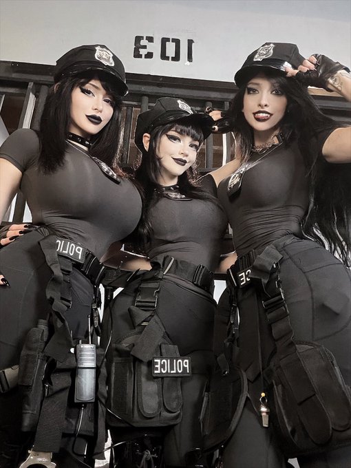 Three women in matching black tactical police-style outfits with caps and gear labeled 'POLICE' pose together in what appears to be a stylized prison or industrial setting.