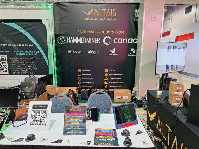 An 'ALTAIR Bitcoin Mining Solutions' trade show booth featuring product banners from Hammerminer, Canaan, Bitmain, ePIC, and Sealminer, with promotional materials and QR codes on the table.