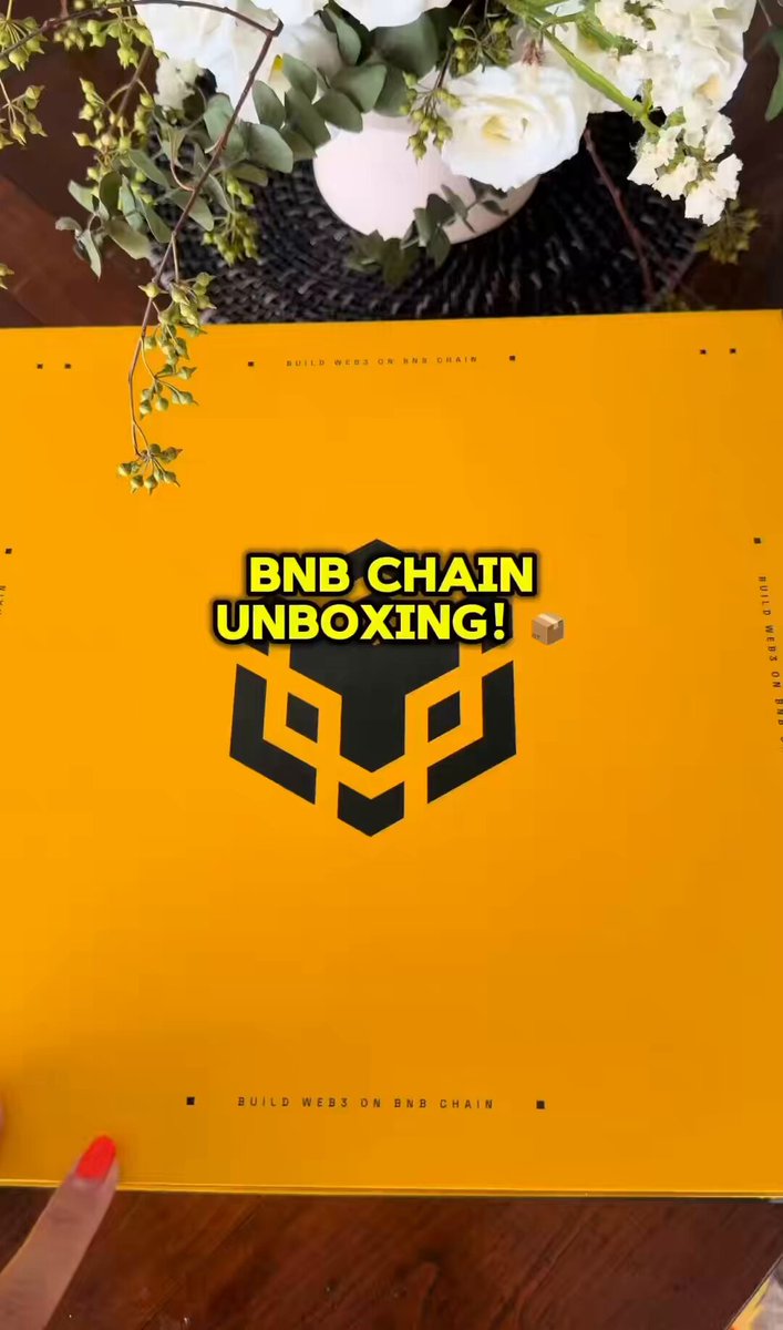 The image features a yellow box with black text that reads "BNB Chain Unboxing!". A person's hand can be seen holding the box, indicating that they are about to open it or have just received it as a gift. The background includes a table with flowers and a vase on top of it, adding an elegant touch to the scene.