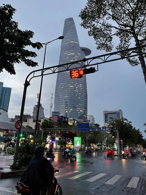 A street view of Ho Chi Minh City featuring the Bitexco Financial Tower in the background, with a traffic light countdown showing 36 seconds and motorcyclists navigating the intersection.
