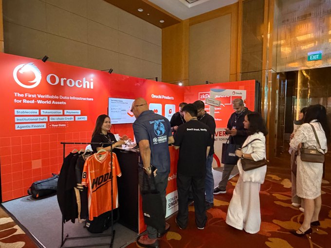 A trade show booth for 'Orochi - The First Verifiable Data Infrastructure for Real-World Assets' with attendees and merchandise including a 'HONA' jersey displayed on a rack.