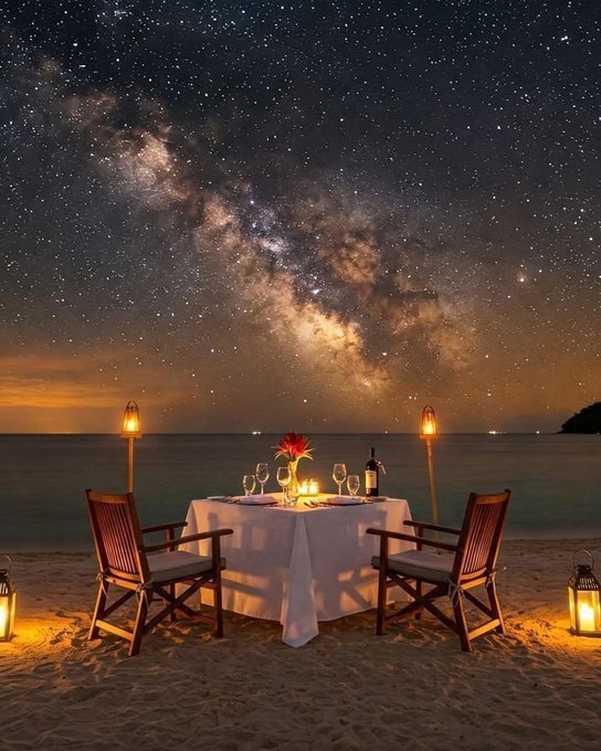 The image features a beautiful beach setting at night, with two chairs placed next to each other facing the ocean. The chairs are positioned under a starry sky, creating an intimate atmosphere for a romantic dinner or date. A dining table is set up nearby, complete with wine glasses and bottles of wine. There's also a vase on the table, adding to the ambiance. 

In addition to the main scene, there are two people visible in the image who appear to be enjoying their time at the beach as well. The overall setting suggests that this could be an outdoor restaurant or a private beachside location where couples can enjoy each other's company and share a romantic meal under the stars.