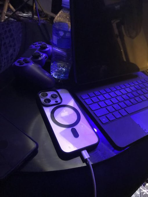 A dimly lit scene showing an iPhone with a MagSafe case charging via cable next to a laptop keyboard, a PlayStation controller, and a water bottle bathed in purple and blue lighting.
