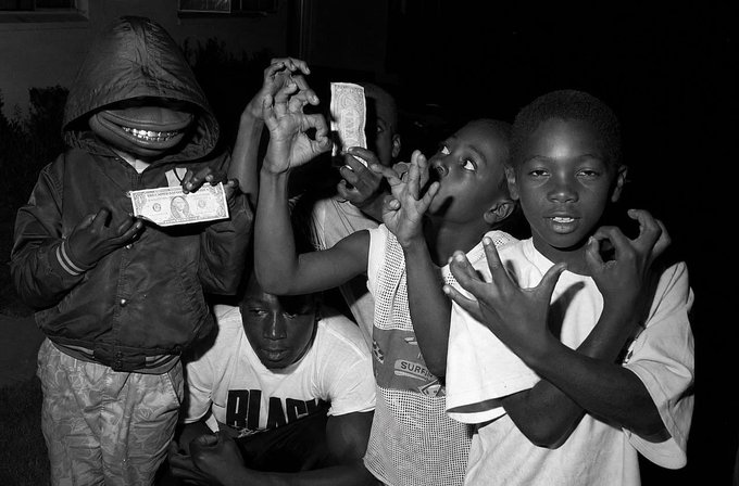 A black and white photograph shows a group of young boys posing at night, with one in a hooded jacket holding a banknote and others making hand gestures, one wearing a 'Superdry' shirt.