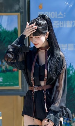 A woman with long dark hair in a high ponytail wears a black sheer blouse with brown harness straps, shielding her eyes with one hand during what appears to be a public event.