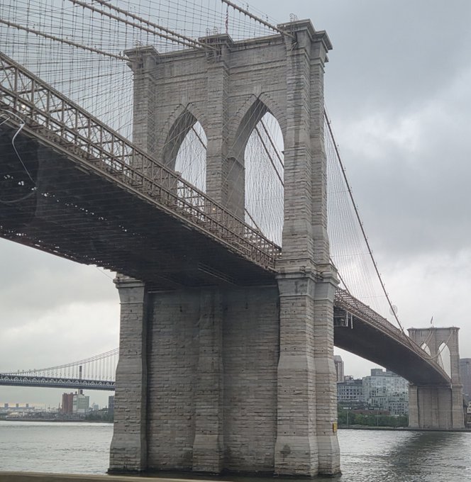 The Brooklyn Bridge stands majestically over the East River on an overcast morning, its iconic stone tower and web of suspension cables framing the iconic NYC skyline in the background—perfectly setting the stage for a trip into the city. In the distance, another bridge peeks through, hinting at the urban adventure awaiting the poster in the "greatest city on Earth."